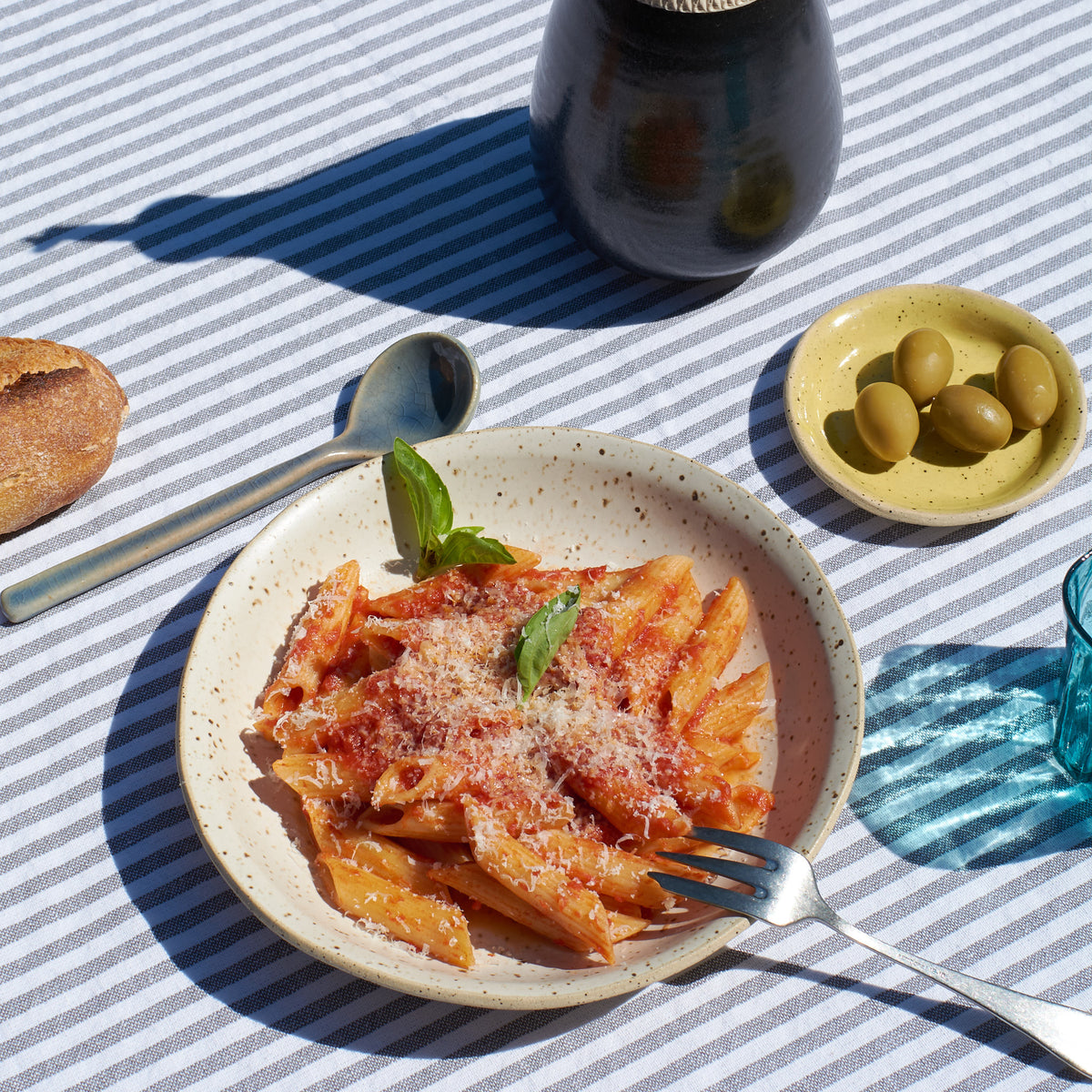 A ceramic speckled pasta bowl by Lola Cera with penne pasta with tomato sauce, grated cheese, and basil garnish on a striped tablecloth, with a spoon, bread, olives, and a dark bottle nearby in bright sunlight.