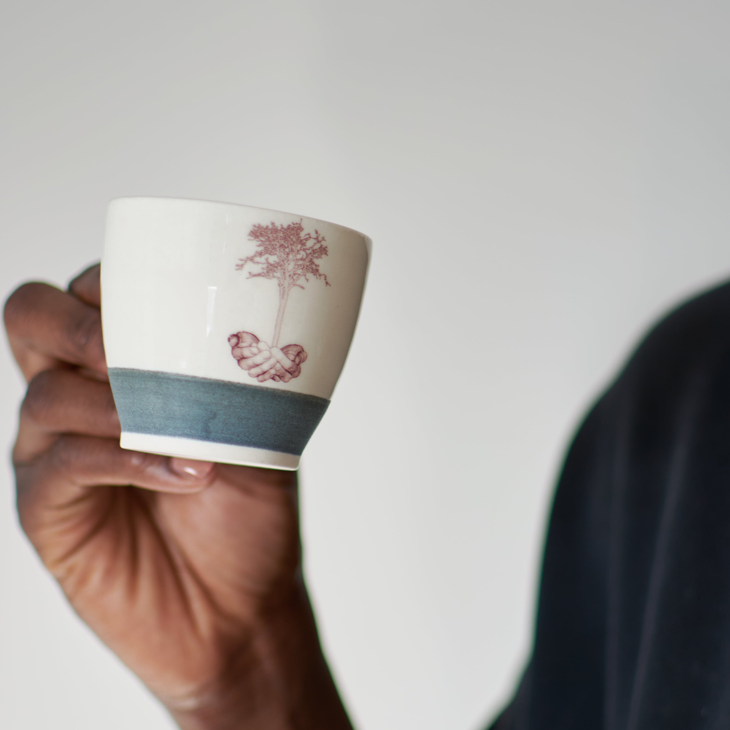 Person holding a Catherine De Abreu espresso cup mug with offering hands and germinating tree decal