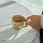 Hand holding a Yukari Hazama spring cherry cappuccino cup on a white tablecloth.