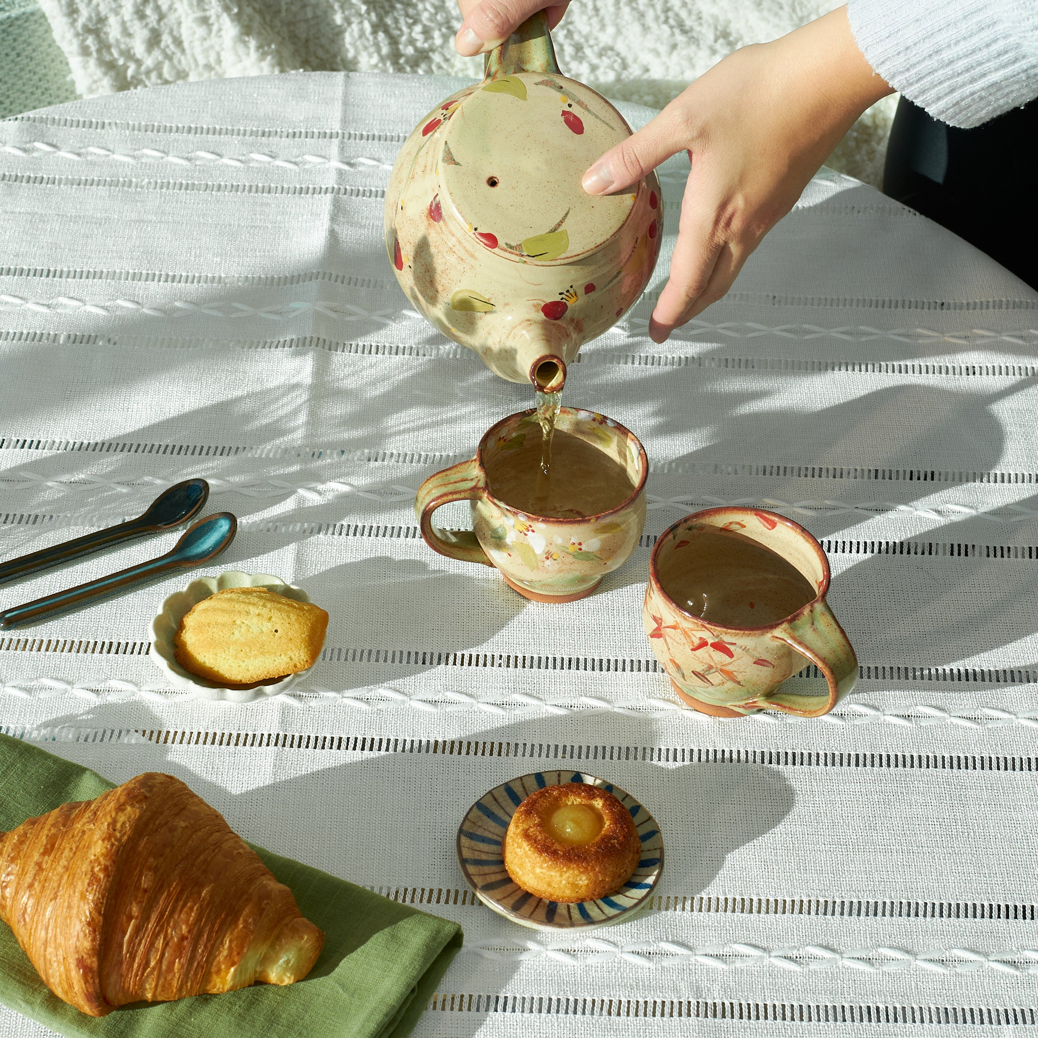 Tea being poured from a Yukari Hazama Iverson teapot into a cup on a white tablecloth with pastries.
